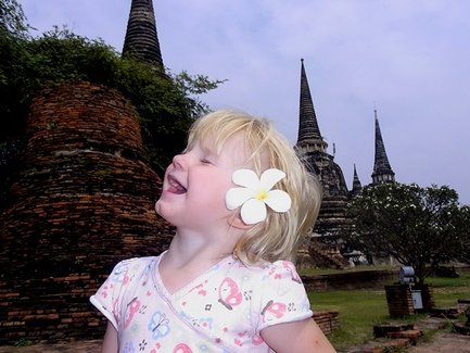Ein fröhliches kleines Mädchen mit blonden Locken lächelt, während sie eine Blume im Haar trägt, vor historischen Tempeln.