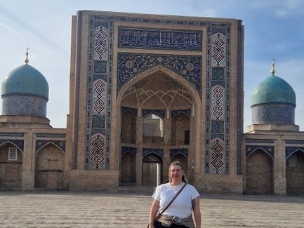 Eine Frau steht auf dem gepflasterten Platz vor dem beeindruckenden Hazrati Imam Komplex mit blauen Kuppeln im Hintergrund.