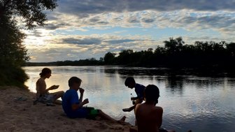 Vier Kinder sitzen am Ufer eines ruhigen Flusses, während die Sonne hinter den Wolken untergeht und den Himmel erleuchtet.