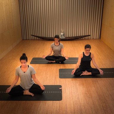 Drei Frauen sitzen in Meditationshaltung auf Yogamatten in einem ruhigen Raum mit Holzfußboden und sanfter Beleuchtung.