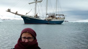 Ein Mann mit einer warmen Mütze und einem dicken Schal steht am Wasser, während ein großes Segelschiff im Hintergrund liegt.