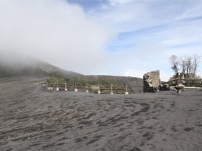 Eine weite, graue Landschaft mit einem sanften Hügel, der von Wolken umhüllt ist, und einem Holzgeländer im Vordergrund.