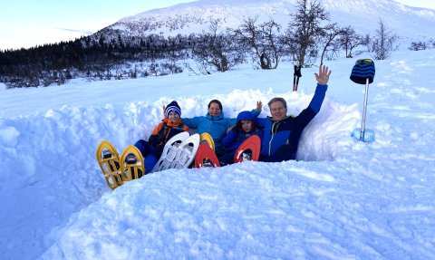 Eine Familie sitzt im Schnee, umgeben von schneebedeckten Bergen, und winkt fröhlich in die Kamera.
