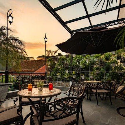 Eine stilvolle Terrasse mit einem Tisch und Stühlen, umgeben von Pflanzen und einem sanften Sonnenuntergang im Hintergrund.