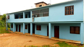 Ein zweigeschossiges, hellblaues Gebäude mit mehreren Fenstern und Türen, umgeben von grünem Gras und einem unbefestigten Weg.