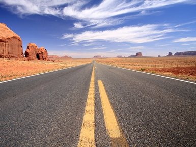 Eine endlose Straße erstreckt sich durch die weite, rote Wüstenlandschaft unter einem strahlend blauen Himmel.