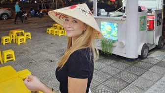 Eine lächelnde Frau trägt einen traditionellen vietnamesischen Hut und steht vor einem Straßenstand mit bunten Stühlen.