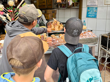Drei Kinder stehen an einem Marktstand und schauen fasziniert auf eine Auswahl an verschiedenen Snacks und Süßigkeiten.