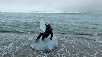 Eine Person sitzt auf einem großen Eisblock am Ufer eines kalten, klaren Gewässers, umgeben von schneebedeckten Bergen.