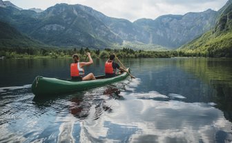 Zwei Personen paddeln in einem grünen Kanu auf einem ruhigen See, umgeben von majestätischen Bergen und üppigem Grün.