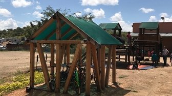 Ein Spielplatz mit einem Holzspielhaus und Fahrrädern, umgeben von einer offenen Landschaft unter einem blauen Himmel.