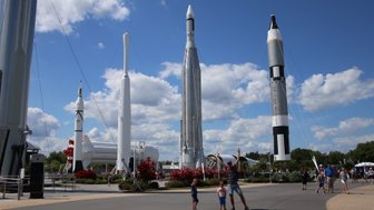 Ein Vater steht mit zwei Kindern vor beeindruckenden Raketen im Kennedy Space Center, umgeben von blühenden Pflanzen und blauem Himmel.