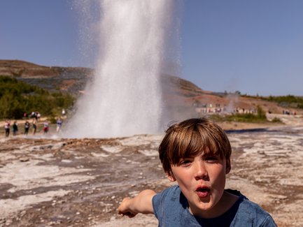 Ein Junge steht vor dem Geysir Strokkur, während er begeistert auf den sprudelnden Wasserstrahl zeigt.
