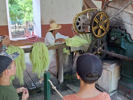 Ein älterer Mann in einem Strohhut arbeitet an einer Maschine, während zwei Kinder aufmerksam zuschauen.