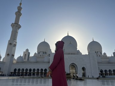 Eine Person in einem roten Gewand steht vor der beeindruckenden Sheikh Zayed Moschee, umgeben von majestätischen Kuppeln.