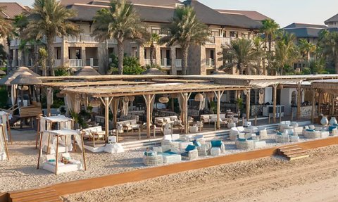 Ein luxuriöser Strandbereich mit gemütlichen Liegen und einem stilvollen Restaurant im Hintergrund, umgeben von Palmen.