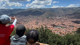 Eine Person zeigt auf die Stadt Cusco, während zwei Kinder fasziniert auf die Aussicht blicken, umgeben von Bergen.