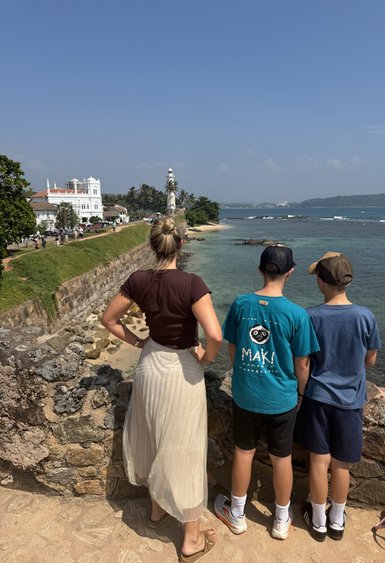 Eine Frau und zwei Kinder stehen am Ufer und blicken auf das Meer, während ein Leuchtturm in der Ferne sichtbar ist.