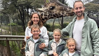 Eine Familie steht auf einer Holzterrasse, während eine Giraffe neugierig in die Kamera schaut und die Kinder lächeln.