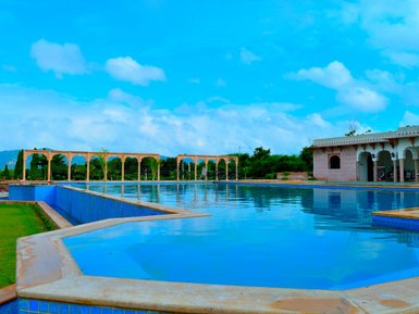 Ein weitläufiger Swimmingpool mit glitzerndem Wasser, umgeben von eleganten Bögen und üppigem Grün unter einem strahlend blauen Himmel.
