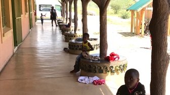 Eine Gruppe von Kindern sitzt auf Baumstamm-Bänken in einem sonnigen Flur, während andere im Hintergrund spielen.