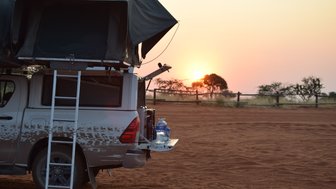 Ein Jeep steht mit aufgebautem Dachzelt auf Sand. Im Hintergrund der Sonnenuntergang - Namibia Familienreise