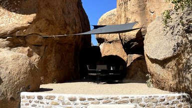 Eine gemütliche Sitzecke unter einem Sonnensegel, umgeben von beeindruckenden Felsen und klarem blauen Himmel.