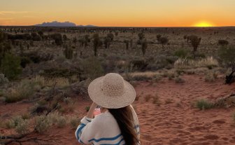Eine Person mit einem Strohhut steht auf rotem Sand und genießt den Sonnenuntergang über der weiten, trockenen Landschaft.