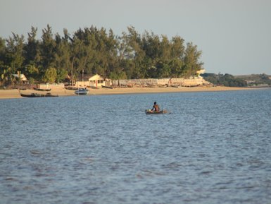 Ein Mann paddelt in einem kleinen Boot auf ruhigem Wasser, während im Hintergrund Bäume und Häuser sichtbar sind.