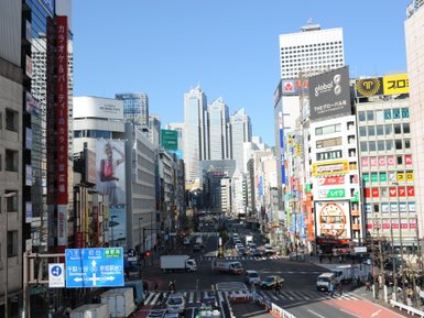 Eine belebte Straße in Tokio mit hohen Gebäuden, bunten Werbetafeln und einem klaren blauen Himmel im Hintergrund.