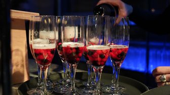 Eine Hand gießt prickelnden Sekt in Gläser, die mit roten und schwarzen Beeren gefüllt sind, auf einem dunklen Tablett.