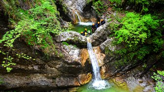 Eine Gruppe von Abenteurern steht an einem kleinen Wasserfall in einer grünen Schlucht, umgeben von üppiger Vegetation.