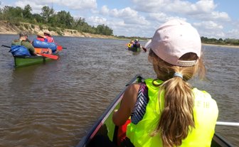 Ein Mädchen in einem gelben Schwimmwesten sitzt im Kanu und beobachtet andere Kanufahrer auf einem ruhigen Fluss.