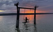 Eine Person schwingt auf einer Holzschaukel im Wasser, während die Sonne hinter einer Wolkendecke untergeht.