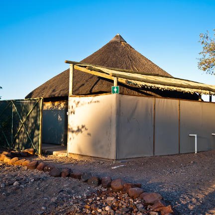 Ein traditionelles Rundhaus mit einem Strohdach steht in der Wüste, umgeben von Bäumen und einem klaren blauen Himmel.
