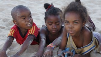 Drei Kinder liegen im Sand, lächeln und spielen, während sie mit ihren Händen im feinen Sand graben.