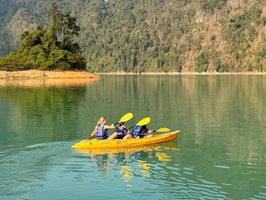 Drei Personen paddeln in einem gelben Kajak auf ruhigem Wasser, umgeben von üppigen Bergen und klarem Himmel.