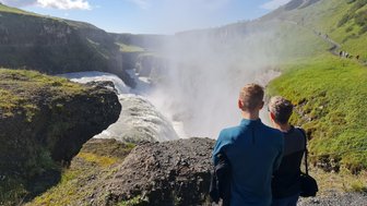 Zwei Personen stehen am Rand des Gullfoss-Wasserfalls und beobachten die tosenden Wassermassen, umgeben von grüner Landschaft.