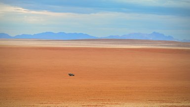 Ein einsames Fahrzeug fährt über die weiten, trockenen Landschaften der Namib-Wüste, vor majestätischen Bergen im Hintergrund.