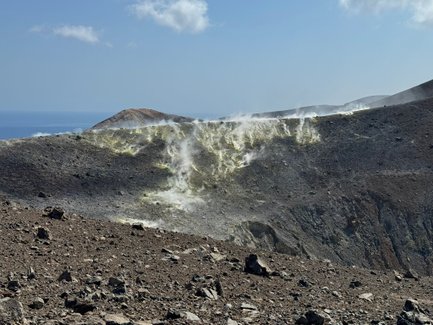 Ein rauchender Vulkan mit einer kargen, steinigen Landschaft und dem blauen Meer im Hintergrund unter einem klaren Himmel.