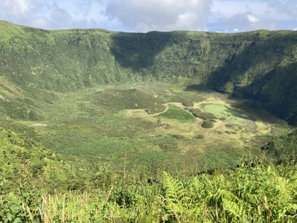 Eine weite, grüne Landschaft mit einem großen Krater, umgeben von steilen, bewaldeten Hängen und sanften Wolken am Himmel.