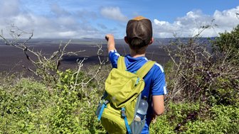 Ein Junge mit einem grünen Rucksack steht auf einem Hügel und zeigt auf die weite Vulkanlandschaft vor ihm.