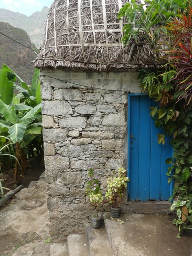 Eine kleine Steinhütte mit einem blauen Holzportal, umgeben von üppigen Pflanzen und einer steinigen Treppe im Freien.