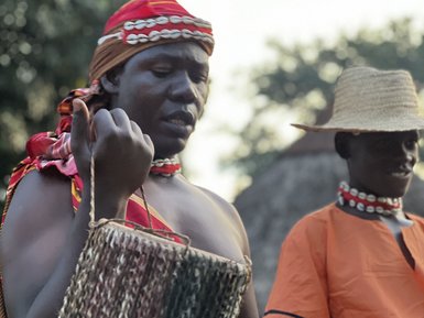 Eine Frau in traditioneller Kleidung spielt ein Musikinstrument, während eine andere Person im Hintergrund zuschaut.