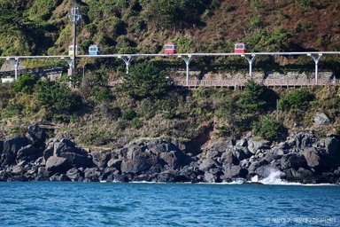 Eine farbenfrohe Seilbahn fährt über eine grüne Küstenlandschaft, während Wellen sanft gegen die Felsen schlagen.