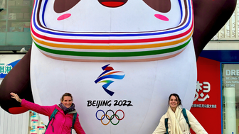 Zwei Frauen posieren fröhlich neben einem großen, aufblasbaren Panda mit den Olympischen Ringen und dem Schriftzug 'Beijing 2022'.
