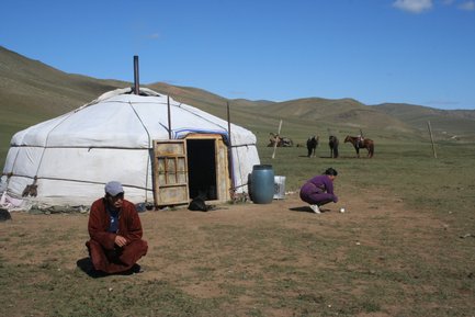 Eine Jurte steht in der Steppe, während ein Mann in traditioneller Kleidung auf dem Boden sitzt und ein anderer arbeitet.