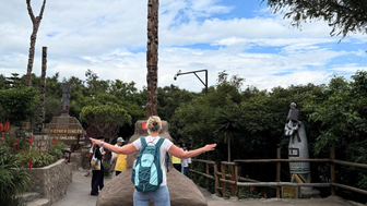 Eine Person mit einem Rucksack steht mit ausgebreiteten Armen vor einem großen, geschnitzten Holzpfahl in einer grünen Umgebung.