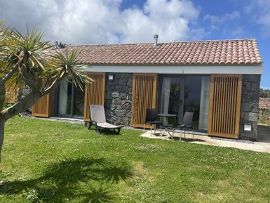 Ein modernes, steinernes Ferienhaus mit Holzjalousien, umgeben von grünem Rasen und einer Palme im Vordergrund.