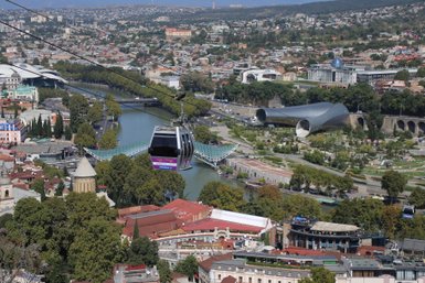 Eine Seilbahn schwebt über die Stadt Tiflis, mit Blick auf den Fluss und moderne Architektur im Hintergrund.